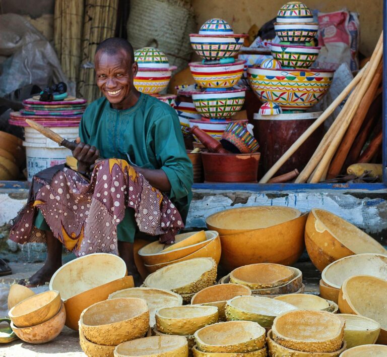 Amid vibrant colors, a smiling artisan sells handmade gourd bowls at an outdoor bazaar.